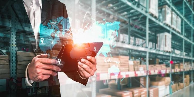 The image shows a businessman in a warehouse looking at an iPad, from which the world map is bursting forth, indicating the use of digital supply chains.