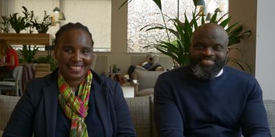 Ester and Tregson sit on a couch in Esther Simpons cafe. They are both smiling, in business attire