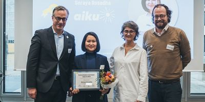 A group of four people (two females and two males) standing in a line, with one female named Soraya proudly holding a certificate for an award win.