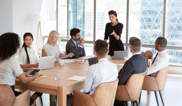 Women and men sat round a boardroom table, with a woman stood chairing the meeting
