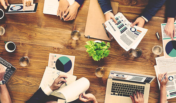 An overhead shot of a meeting featuring people using laptops, holding paperwork and drinking coffee