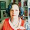 White middle aged woman in red dress with tattooed arms and green short curly hair, in front of bookcases