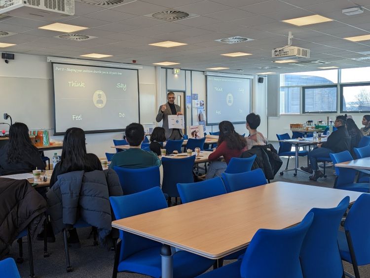 Rashik leads the seminar and is stood at the front explaining the topic of his talk. All the students are captivated. 
