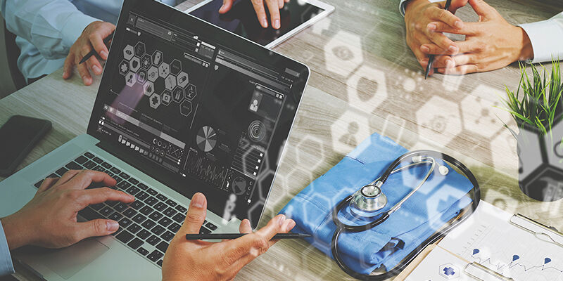 A desk shot of people using laptop and tablet, with doctor's equipment on the table