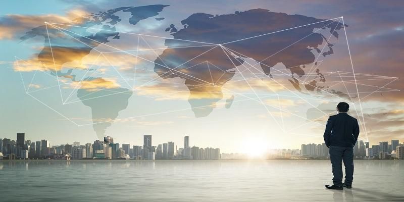 The picture shows the back of a professional man standing on the right-hand side of the image, looking at a city skyline, while in the sky a map of the world is displayed with lines joining the various countries, thus indicating international business.